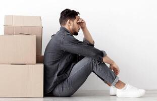 Frustrated arab couple sitting by paper boxes on floor in empty apartment, having problem with relocating, white wall background, side view, copy space. Depressed middle-eastern family at new home photo
