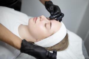 A woman lies on a treatment table while a beauty technician applies permanent makeup to her eyebrows. The salon environment features a calming atmosphere as the procedure enhances her features. photo