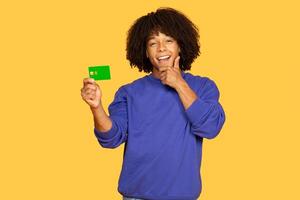 A young black man with curly hair is smiling while holding a green card, showing a thoughtful expression. The vibrant yellow background adds energy to the moment, emphasizing his happiness. photo