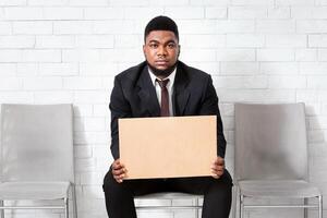 African American man in formal suit holding LOOKING FOR JOB sign, waiting for work interview at office lobby, empty space. Millennial black guy seeking permanent employment at big company photo