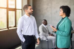 Two professionals are talking in a bright office space, showcasing teamwork and diversity. Others are seated in the background, highlighting collaboration in a corporate environment. photo