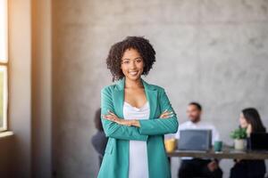 In a contemporary office featuring large windows, a confident woman stands with crossed arms. Behind her, a diverse team engages in discussion by a table, highlighting teamwork and collaboration. photo