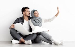 Emotional muslim couple thinking about design in their new house, sitting on floor over white empty wall, holding apartment plan and imaging future interior, panorama with copy space photo