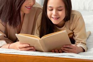 Family stay home hobbies. Lovely young mother and her teen daughter reading book together on bed indoors. Cute girl and her mom enjoying interesting story during covid-19 lockdown photo
