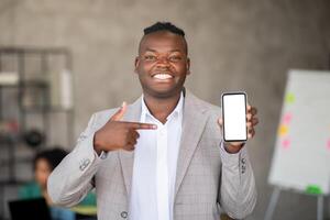 In a contemporary office, a smiling man points to his smartphone, highlighting its features. Team members collaborate in the background, reflecting a dynamic work environment focused on success. photo
