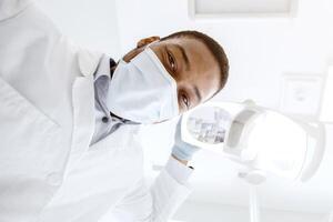 Low Angle View Of Black Stomatologist Turning On Lamp Before Check Up With Patient, Professional African American Dentist Doctor In Medical Mask Looking At Camera, Ready For Making Treatment photo