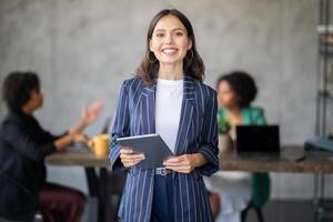 A diverse group of professionals collaborates in a modern office. One woman stands in front, confidently holding a tablet as colleagues discuss strategies and ideas, fostering teamwork and success. photo