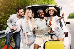 Hitchhiking concept. Group of young friends looking at map during road trip, taking selfie while sitting in car trunk on highway, copy space. Diverse students resting on roadside after long drive photo