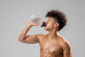 A young Brazilian man with an athletic build is quenching his thirst with a water bottle. He is in a gym setting, highlighting a commitment to a healthy lifestyle and fitness. photo