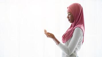 Portrait of young black muslim woman praying near window at home, religious lady in headscarf standing with closed eyes, making islamic pray dua gesture with cupped hands, side view, copy space photo