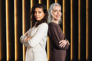 Two confident business leaders pose back to back in a stylish office environment. The focus is on their professional demeanor and strong presence amidst contemporary backdrop. photo