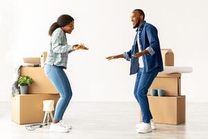 Happy Young Black Spouses Dancing In Their New Flat On Moving Day, Cheerful African American Couple Having Fun Among Cardboard Boxes With Belongings While Relocating To Own Apartment, Copy Space photo