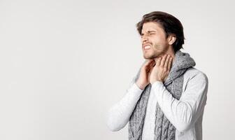 Sore throat, inflammation of tonsils. Man in scarf suffers from pain and touches his throat, isolated on gray background, studio shot photo
