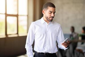 A business professional checks his smartphone in a contemporary office environment. The warm lighting and diverse team in the background enhance the atmosphere of collaboration and productivity. photo