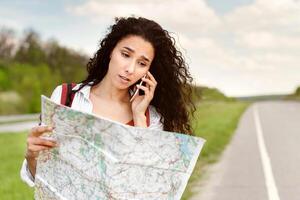 Stressed female tourist with map standing on roadside, speaking on smartphone, cannot catch car or find her travel route. Young lady feeling lost while hitchhiking on highway, having problem photo