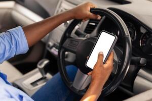Over the shoulder view of happy black male driver holding smartphone with white blank screen, using mobile app for navigation, mock up. Closeup of cell phone with empty display, copy space template photo