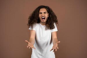 A young European man with curly, long hair appears passionately expressive while standing in a studio. The neutral background contrasts with his vibrant energy and animated gestures. photo