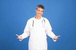 A mature Caucasian man in a white doctor coat stands confidently with a welcoming smile. He gestures with open arms, promoting a friendly atmosphere in a healthcare setting. photo