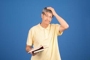 A middle aged Caucasian man in a yellow shirt appears confused while holding an open book. The plain blue background emphasizes his expression of uncertainty and contemplation. photo