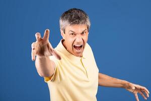 A middle aged man in his 50s is passionately expressing intense emotion. He appears to be in a studio with a blue background, wearing a yellow shirt and with his hand extended forward. photo