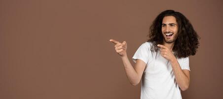 A young Caucasian man with long, curly hair joyfully poses while pointing at copy space. His carefree expression reflects a vibrant and playful attitude toward life. photo