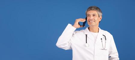 A mature Caucasian man, dressed in a white coat, stands against a blue background while talking on a smartphone. His expression shows engagement, copy space photo
