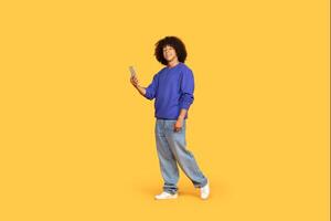 A young black man smiles while standing in relaxed jeans and a sweater, confidently engaging with his smartphone. The vibrant yellow background creates a cheerful atmosphere. photo