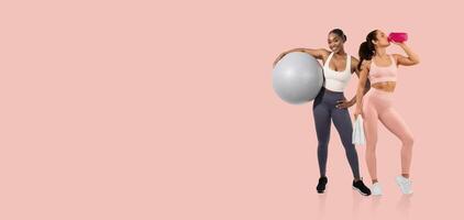 Two women are engaged in an active workout session in a fitness studio. One holds a large exercise ball, while the other drinks from a sports bottle, showcasing a healthy lifestyle. photo