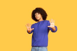 A Brazilian guy stands against a bright yellow backdrop, wearing a blue sweater and jeans. He strikes a playful pose with arms raised, showcasing a confident and relaxed vibe. photo