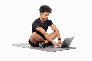 A young Brazilian man sits on a gym mat, balancing fitness and technology. He is focused on his laptop, blending a healthy lifestyle with digital engagement in a bright, isolated space. photo