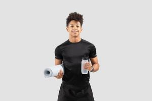 A young Brazilian man stands confidently, holding a gym mat and a water bottle. He embodies an active lifestyle, showcasing his athletic build and fitness enthusiasm in a bright, isolated setting. photo