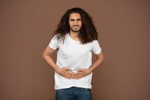 A young Caucasian man with long, curly hair is casually standing against a brown backdrop. He is holding his stomach, have pain photo