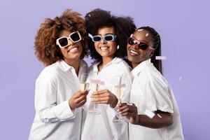 Three young African American women, striking joyful poses with sparkling drinks, showcase their friendship. They wear stylish sunglasses and white shirts against a purple background. photo