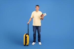 A middle aged European man stands in front of a blue backdrop, holding a suitcase and travel documents. He appears cheerful and ready for an upcoming adventure in a carefree setting. photo
