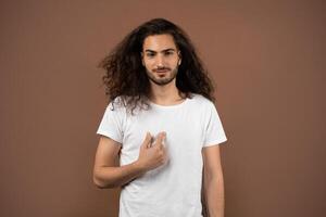 A young man with long, curly hair stands against a brown backdrop. He appears relaxed and confident, pointing toward himself with a slight smile. photo