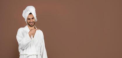 A young man with curly hair is smiling and pointing playfully while wearing a white bathrobe and a towel on his head. The background features a warm brown tone creating a cozy atmosphere, copy space photo