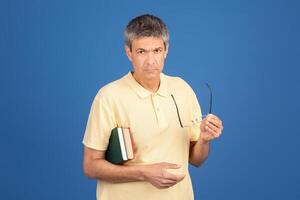 A middle aged man in his 50s stands against a blue background. He holds glasses in one hand and books in the other, looking contemplative and slightly isolated in his surroundings. photo