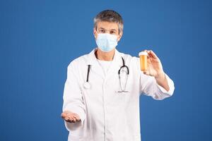 A middle aged Caucasian man, dressed in a white coat and mask, displays a prescription bottle while extending his other hand. He appears focused and ready to assist in a healthcare environment. photo