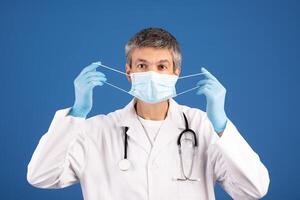 A mature Caucasian man in a white lab coat puts on a face mask, ready to engage in patient care. He wears gloves and has a stethoscope around his neck, indicating a commitment to health. photo