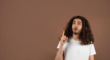 A young Caucasian man with dark, curly hair expresses thoughtful curiosity while pointing upwards at copy space. He is styled casually in a simple white shirt against a brown backdrop. photo