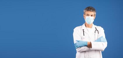 A middle aged Caucasian man dressed in a white lab coat and gloves stands with his arms crossed. He wears a surgical mask, emphasizing his role in healthcare, copy space photo