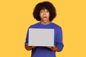 A surprised young man holds a laptop in front of him, showcasing his expressive reaction. He has curly hair and wears a blue sweater, creating a vivid contrast with the yellow background. photo