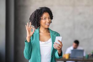 A team member in a corporate office smiles while holding a smartphone and giving an okay gesture. Collaboration thrives in this diverse environment, showcasing successful teamwork. photo