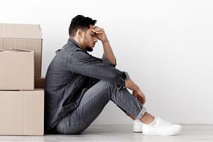 Upset middle-eastern family sitting separated by boxes on floor in empty apartment, having problem with relocating, white wall background, side view, copy space. Frustrated arabic couple at new home photo