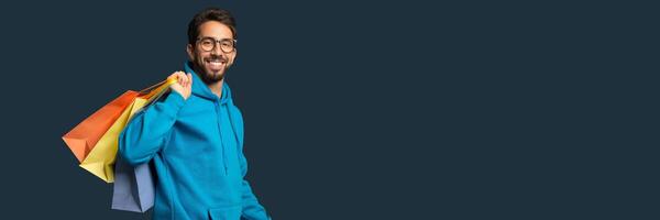Shopping Concept. Handsome Smiling Young Man Holding Colorful Paper Shopper Bags, Happy Millennial Guy In Eyeglasses Carrying Purchases, Standing Against Blue Studio Background, Free Space photo