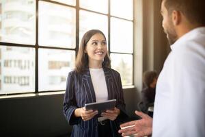 Two professionals are interacting in a bright office space. One person is holding a tablet, while the other is speaking animatedly. Their teamwork reflects a commitment to success and diversity. photo