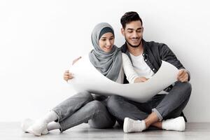 Cheerful middle-eastern family husband and wife in hijab sitting on floor in empty room, holding apartment scheme, copy space for advertising, white blank wall background, panorama photo