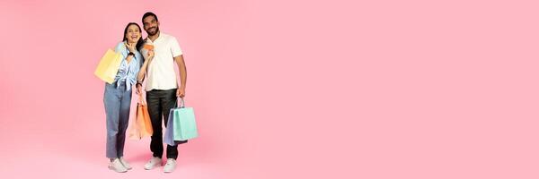 Happy cheerful beautiful young couple shopping together on pink studio background, handsome guy and pretty lady holding colorful paper bags, purchases and bank card, copy space photo