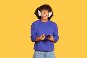 A young black man with curly hair smiles while holding a game controller. He is wearing headphones and is immersed in gaming, showcasing a lively atmosphere in a bright yellow space. photo