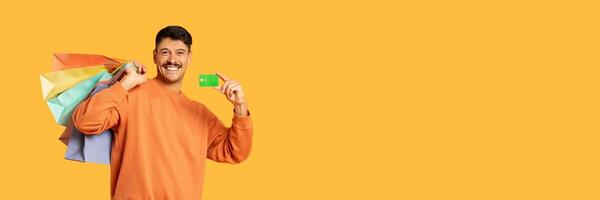 Smiling man carrying colorful shopping bags and showing a credit card, on a plain yellow background, copy space photo